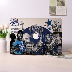 May include: A laptop with a sticker design featuring the words "UTOPIA" and "KEEP FASHION WEIRD". The design includes images of celebrities, money, and other graphics. The laptop is on a white surface.