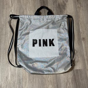 Puede incluir: Mochila plateada con cordón, correas negras y asa negra. La palabra "PINK" está impresa en letras negras en un cuadrado blanco en la parte delantera de la bolsa. La bolsa tiene un aspecto brillante y brillante.