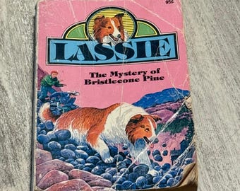 May include: A vintage paperback book titled "Lassie: The Mystery of Bristlecone Pine." The cover features a collie dog, a boy, and a mountain scene. The book has a pink cover with the title "Lassie" in large blue letters.