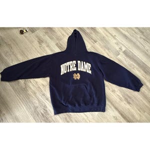 Puede incluir: Sudadera con capucha azul marino con el texto "NOTRE DAME" en letras blancas en el pecho. Debajo, un logotipo dorado y azul marino. La sudadera tiene un bolsillo delantero y una capucha.