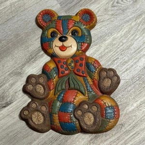 May include: A colorful, patchwork-style teddy bear wall hanging. The bear features a variety of colors including red, blue, yellow, and brown. It has a red bow tie with white polka dots and detailed paw pads. The bear is a decorative item.