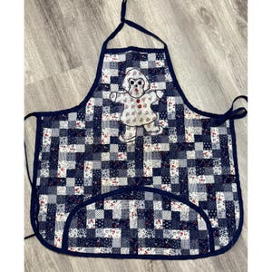 Puede incluir: Delantal acolchado con diseño patchwork en tonos azules, blancos y rojos. Presenta una aplicación de hombre de jengibre. Ribete y lazos azul oscuro. Ideal para la cocina.
