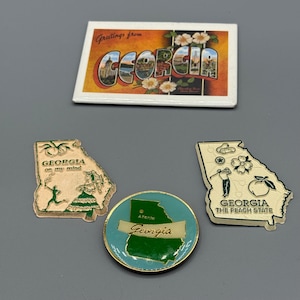 May include: A collection of Georgia-themed souvenirs, including a postcard with "Greetings from Georgia" and images of landmarks, and three pins in the shape of the state. One pin says "Georgia on my mind", another says "Atlanta Georgia", and the last one says "Georgia The Peach State".