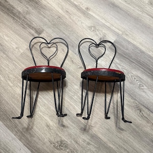 Puede incluir: Dos pequeñas sillas de metal negro con respaldos en forma de corazón. Los asientos redondos tienen cojines rojos y bases de madera. Las sillas tienen patas finas y curvas.