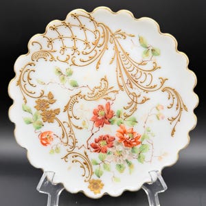 May include: A white decorative plate with a scalloped edge and gold trim. The plate features hand-painted floral designs in red, orange, and white, with gold filigree accents. The plate is displayed on a clear stand.