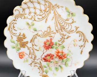 May include: A white decorative plate with a scalloped edge and gold trim. The plate features hand-painted floral designs in red, orange, and white, with gold filigree accents. The plate is displayed on a clear stand.