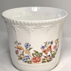 May include: White ceramic planter with a floral and butterfly design. The planter has a scalloped rim and embossed details. The floral design includes red, blue, yellow, and pink flowers, with a butterfly.
