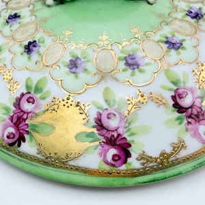 May include: A round porcelain lid featuring a light green border and a white center decorated with hand-painted pink and purple rose clusters, green leaves, and intricate gold filigree scrollwork accents throughout the surface.