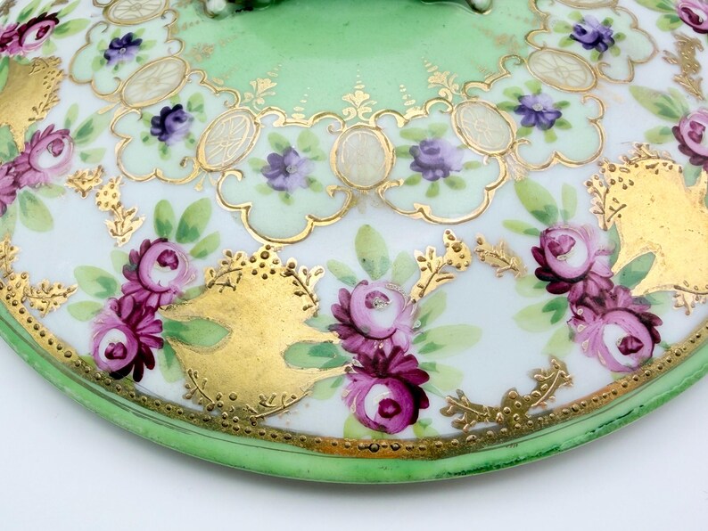 May include: A round porcelain lid featuring a light green border and a white center decorated with hand-painted pink and purple rose clusters, green leaves, and intricate gold filigree scrollwork accents throughout the surface.