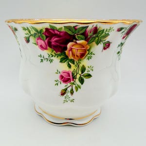 May include: A white porcelain planter featuring a scalloped rim and base with gold trim. The exterior is decorated with a floral pattern of deep red, pink, and yellow roses with green leaves and stems arranged in a cascading design around the body.