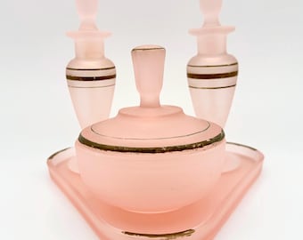 May include: A set of three pieces of frosted pink glass vanity items consisting of two tall bottles with stoppers and one lidded jar, all arranged on a matching triangular tray. Each piece features thin, horizontal gold-toned painted bands.