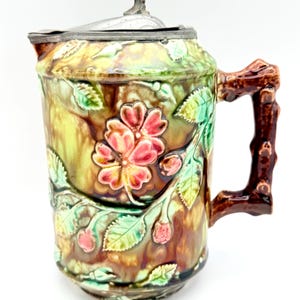 May include: A vintage ceramic pitcher with a detailed floral design. The pitcher features a brown, green, and yellow color scheme with a silver-toned lid and a branch-like handle. The floral pattern includes pink flowers and green leaves.