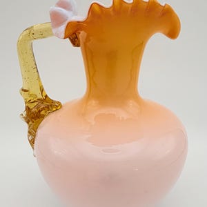May include: A decorative glass pitcher with a rounded body and a ruffled rim. The pitcher is primarily a soft pink color, transitioning to a warm orange at the top. It has a yellow handle with floral accents.