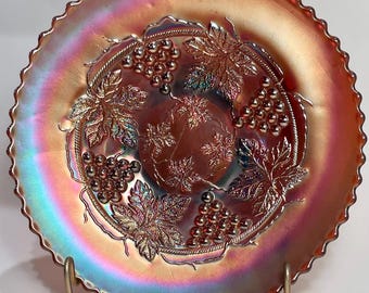 May include: Iridescent glass plate with a grape and leaf design. The plate has a ruffled edge and is supported by a gold-colored metal stand. The colors shift from orange to purple and green.