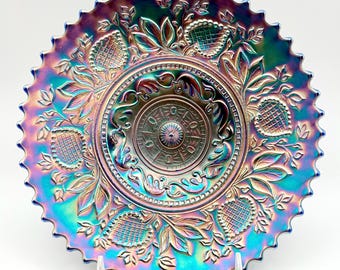 May include: A round iridescent carnival glass plate featuring a scalloped edge and a central medallion design. The surface displays a vibrant spectrum of blue, purple, and gold tones with embossed patterns of stylized leaves and textured fruit motifs.