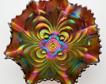 May include: A round glass bowl featuring an iridescent carnival finish with a vibrant spectrum of red, gold, green, and blue hues. The surface displays a raised, symmetrical floral pattern with central concentric circles and scalloped edges.
