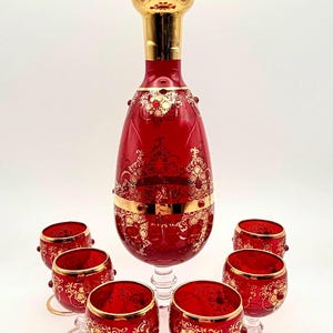 May include: A red glass decanter with a gold stopper and six matching red glass goblets. Each piece features intricate gold filigree patterns, gold rim accents, and small red glass bead embellishments on the exterior surfaces.