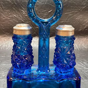 May include: A vintage cobalt blue glass salt and pepper shaker set. The set includes two shakers with silver-colored lids and a matching handle. The shakers and handle are connected by a rectangular base. The glass has a textured, diamond-like pattern.