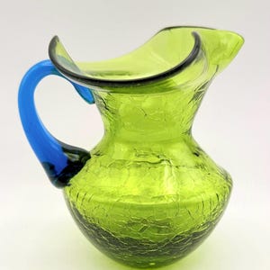 May include: A small, green glass pitcher with a crackle finish. The pitcher has a blue handle and a black rim. The pitcher is likely used for serving beverages and has a unique, decorative appearance.