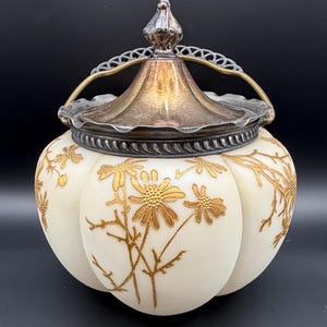 May include: An antique white jar with a silver lid and handle. The jar is decorated with gold floral designs. The lid has a decorative silver handle and finial. The jar is likely made of glass or porcelain.