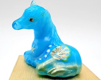 May include: A bright blue glass figurine of a reclining horse. The horse features a white star-like pattern on its chest, a row of white dots along its back, and a painted shell-like design on its flank with gold-toned glitter accents.