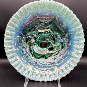 May include: Iridescent green and blue glass plate with a ruffled edge. The plate features a detailed embossed design with a central floral motif and surrounding figures. The glass has a shimmering, opalescent finish.