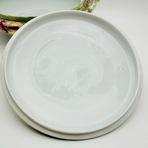 May include: A white porcelain tureen lid with a smooth, circular surface, positioned next to a matching tureen base featuring a green rim, gold accents, and a floral pattern with pink roses and green leaves on the exterior side.