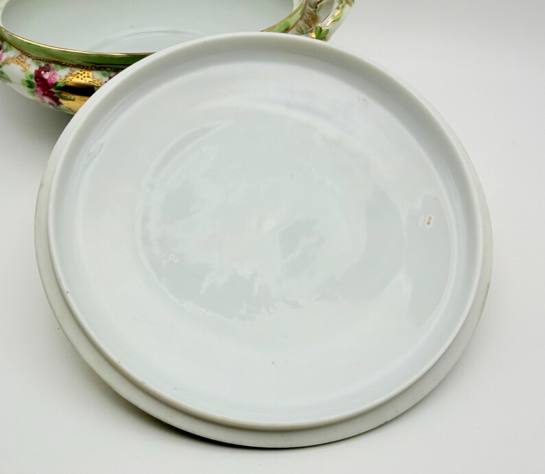 May include: A white porcelain tureen lid with a smooth, circular surface, positioned next to a matching tureen base featuring a green rim, gold accents, and a floral pattern with pink roses and green leaves on the exterior side.