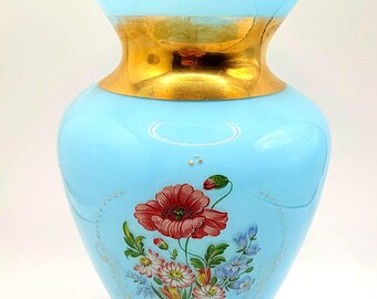 May include: A light blue glass vase with a gold rim and base. The vase features a hand-painted floral design with a large red poppy and other colorful flowers. The vase has a ruffled top.