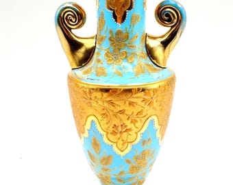 May include: An ornate, antique vase with a gold finish and a light blue central band. The vase features intricate floral designs and two gold handles. The base and rim are also gold, creating a striking contrast.