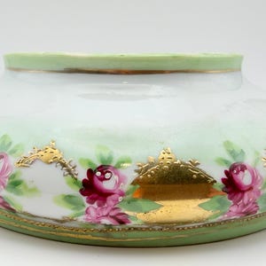 May include: A low, wide porcelain bowl featuring a pale green rim and base. The white body is decorated with hand-painted pink roses, green leaves, and ornate gold leaf accents arranged in a repeating pattern around the exterior surface.