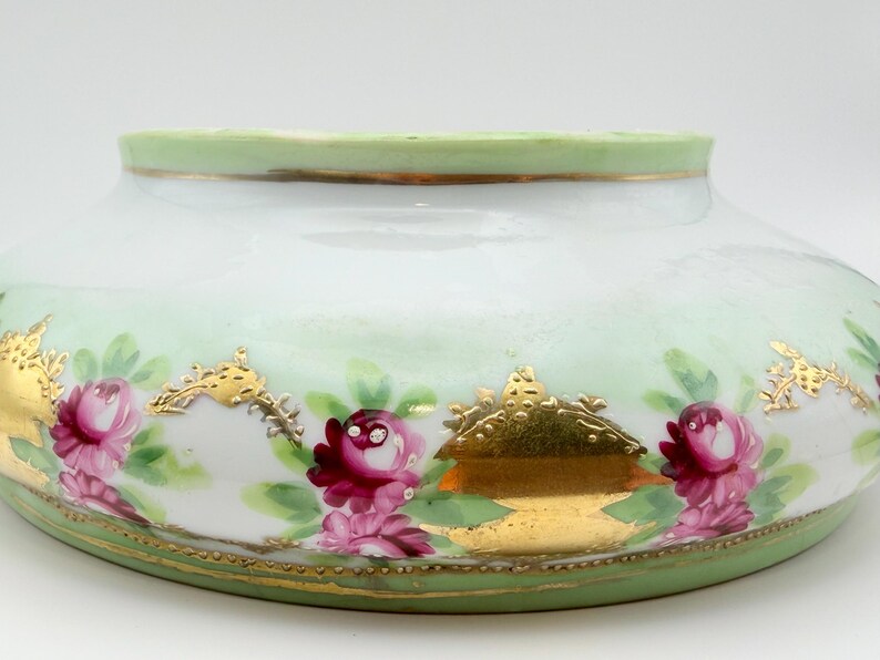 May include: A low, wide porcelain bowl featuring a pale green rim and base. The white body is decorated with hand-painted pink roses, green leaves, and ornate gold leaf accents arranged in a repeating pattern around the exterior surface.