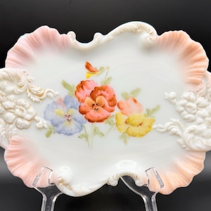 May include: White porcelain serving dish with a scalloped edge in pink. The dish features a hand-painted floral design with orange, yellow, and blue pansies. Raised floral relief details are on the sides.
