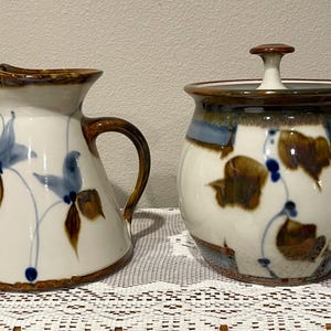 May include: A ceramic pitcher and lidded jar set. The pitcher has a brown handle and spout, with blue and brown floral designs on a white background. The jar has a matching design and a brown lid with a small knob. The set is on a white lace tablecloth.