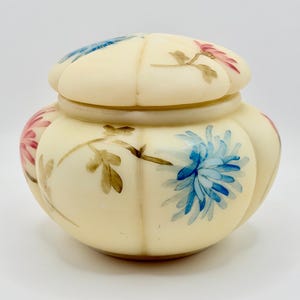 May include: Cream-colored, lidded jar with a floral design. The jar features a rounded shape with vertical ridges and is adorned with hand-painted blue, pink, and brown flowers. The lid mirrors the jar's shape and is also decorated with flowers.