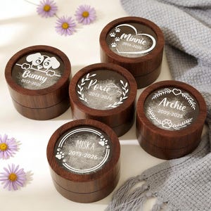 May include: Five round wooden pet keepsake boxes with clear tops. Each box has a different pet's name and dates printed in white. The boxes are arranged on a white surface with purple flowers.