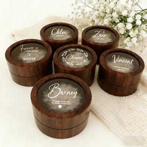 May include: Six dark brown wooden keepsake boxes with clear lids, each inscribed with a name and dates. The boxes have a wood grain effect. Some boxes feature the text "Forever in our heart". The boxes are displayed on a white surface.