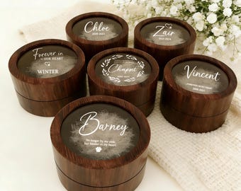 May include: Six wooden keepsake boxes with clear tops, each engraved with a name and dates. The boxes are dark brown with a wood grain finish. Some boxes have text that reads "Forever in our heart". The boxes are arranged on a white surface.