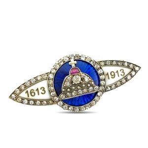 May include: A silver brooch featuring a central circular blue enamel medallion with a diamond-encrusted crown and a red stone. The brooch is flanked by two white enamel wings displaying the years 1613 and 1913, all bordered by small clear stones.