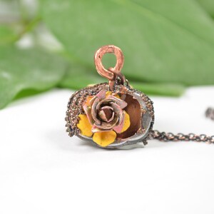 May include: A delicate copper and silver necklace with a yellow and pink flower pendant. The pendant is made of metal and has a textured surface. The necklace is on a white background.