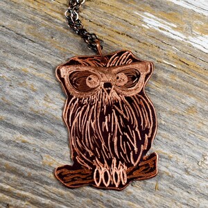 以下が含まれることがあります： メガネをかけた銅色のフクロウのペンダント。銀色のチェーンに吊り下げられています。フクロウはエッチングされた線で詳細に描かれており、ペンダントは風化した木製の背景に配置されています。フクロウは枝にとまっています。