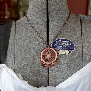 May include: A copper necklace with a pendant featuring a bicycle wheel design and the words "True Love". The necklace is on a grey dress form with a blue and white label that reads "Acme Adjustable Dress Form".