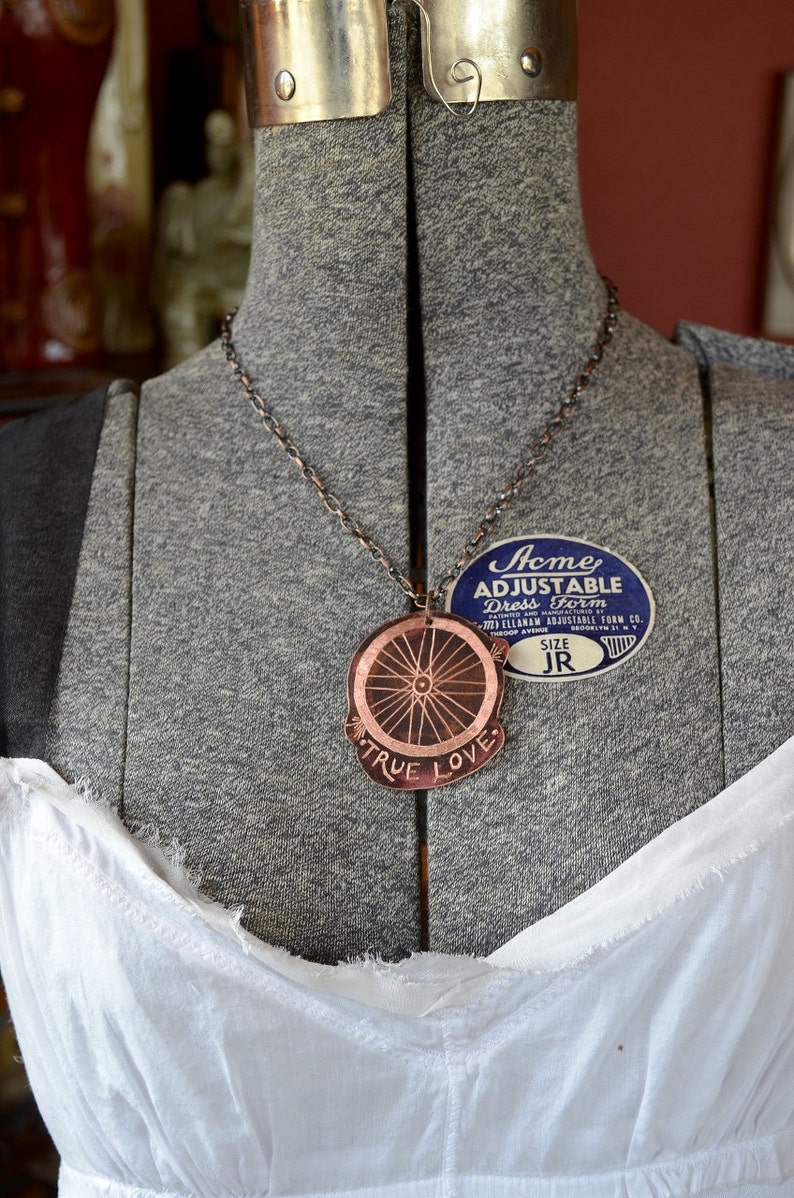 May include: A copper necklace with a pendant featuring a bicycle wheel design and the words "True Love". The necklace is on a grey dress form with a blue and white label that reads "Acme Adjustable Dress Form".