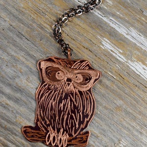 以下が含まれることがあります： 詳細なエッチングデザインが施された銅製のフクロウペンダントネックレス。フクロウのペンダントは、暗いチェーンから吊り下げられています。ペンダントはメガネをかけたフクロウが特徴です。ネックレスは木製の表面に表示されています。