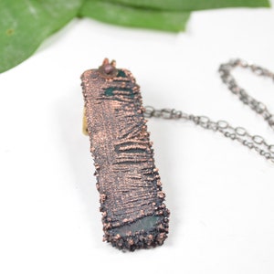 May include: A copper and black necklace with a textured rectangular pendant. The pendant has a rough, organic texture and a dark green patina.