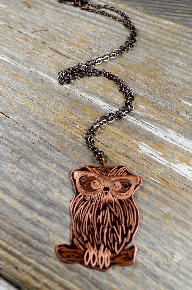 以下が含まれることがあります： 銅色のフクロウペンダントネックレス、暗いチェーン付き。 フクロウのペンダントは、メガネと質感のある羽を備えた詳細なデザインが特徴です。 ネックレスは、風化した木製の表面に表示されています。