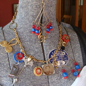 Op de afbeelding: Een gouden ketting met verschillende bedels, waaronder een noot, een bloem, een speld en een klein fotolijstje. De ketting heeft ook rode en blauwe kralen.