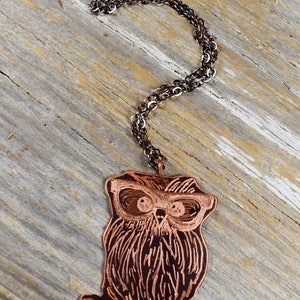 以下が含まれることがあります： 銅色のフクロウペンダントネックレス、暗いチェーン付き。 フクロウのペンダントは、エッチングされた線とメガネで詳細に描かれています。 ネックレスは、風化した木製の表面に表示されています。