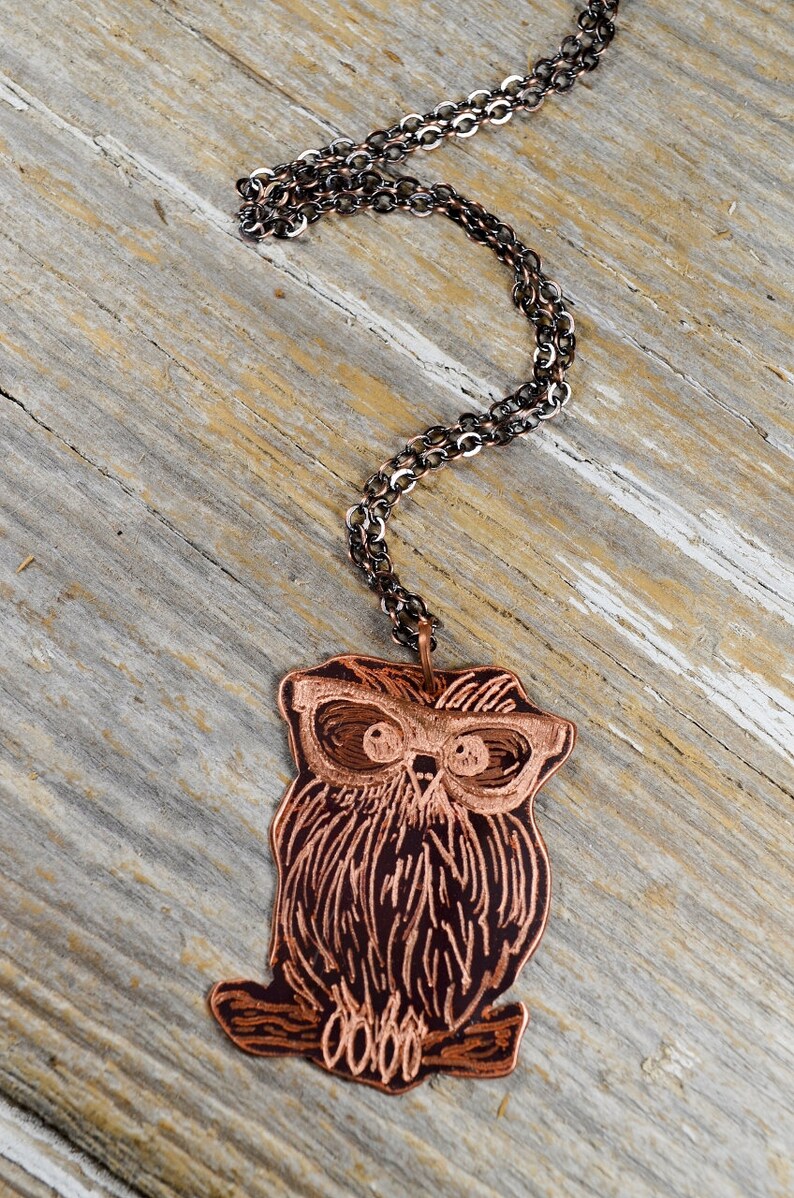 以下が含まれることがあります： 銅色のフクロウペンダントネックレス、暗いチェーン付き。 フクロウのペンダントは、エッチングされた線とメガネで詳細に描かれています。 ネックレスは、風化した木製の表面に表示されています。