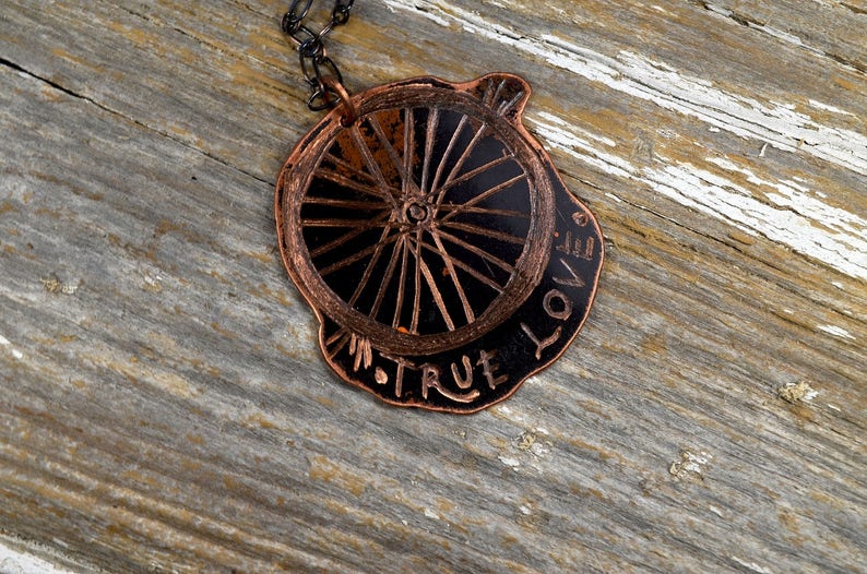 May include: A copper necklace featuring a wheel-shaped pendant with the words "TRUE LOVE" inscribed around the edge.